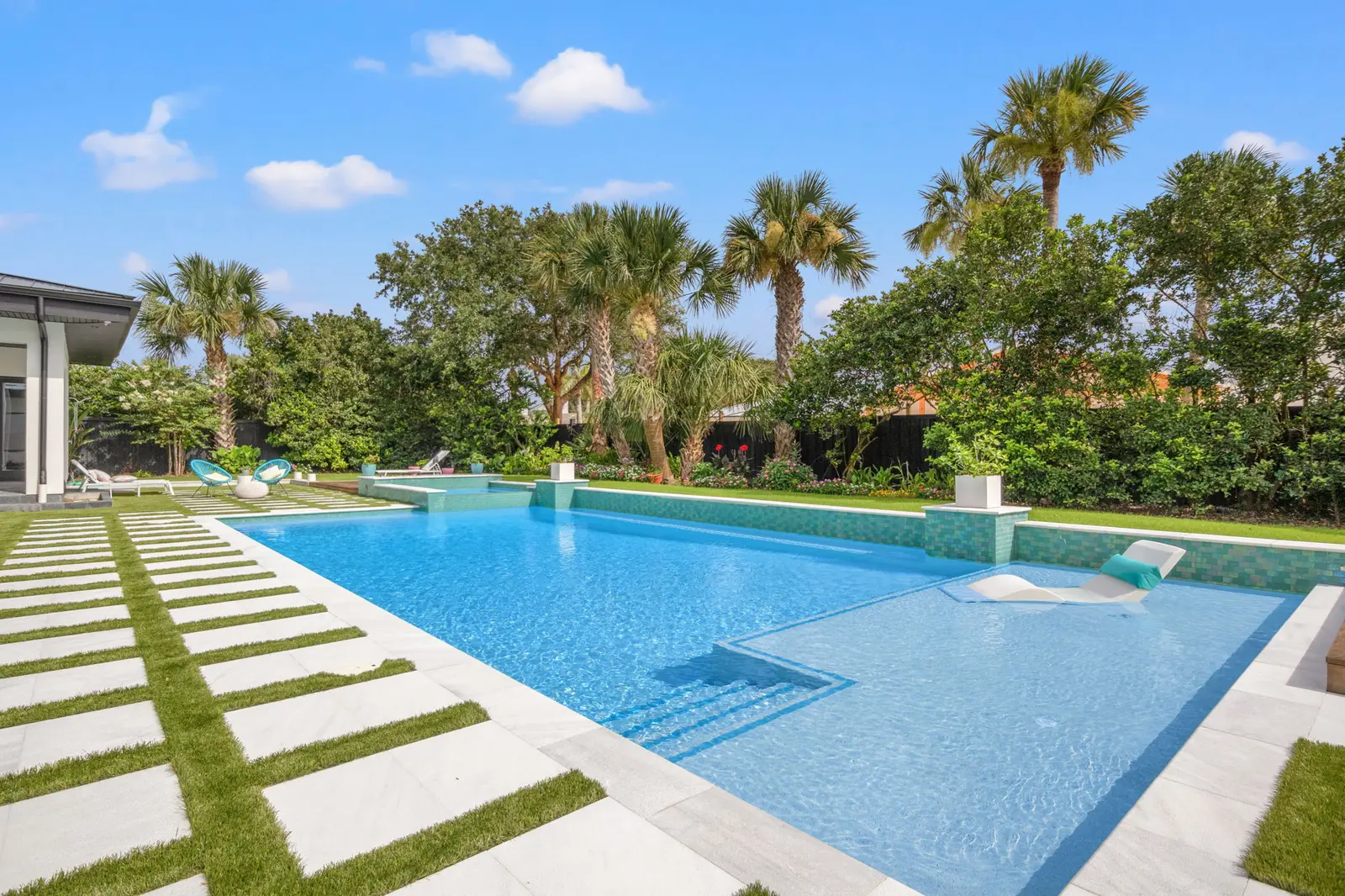 Sparkling blue luxury pool with geometric white deck, manicured lawn, and palm trees under clear sky - Coastal Luxury Outdoors, Jacksonville, FL