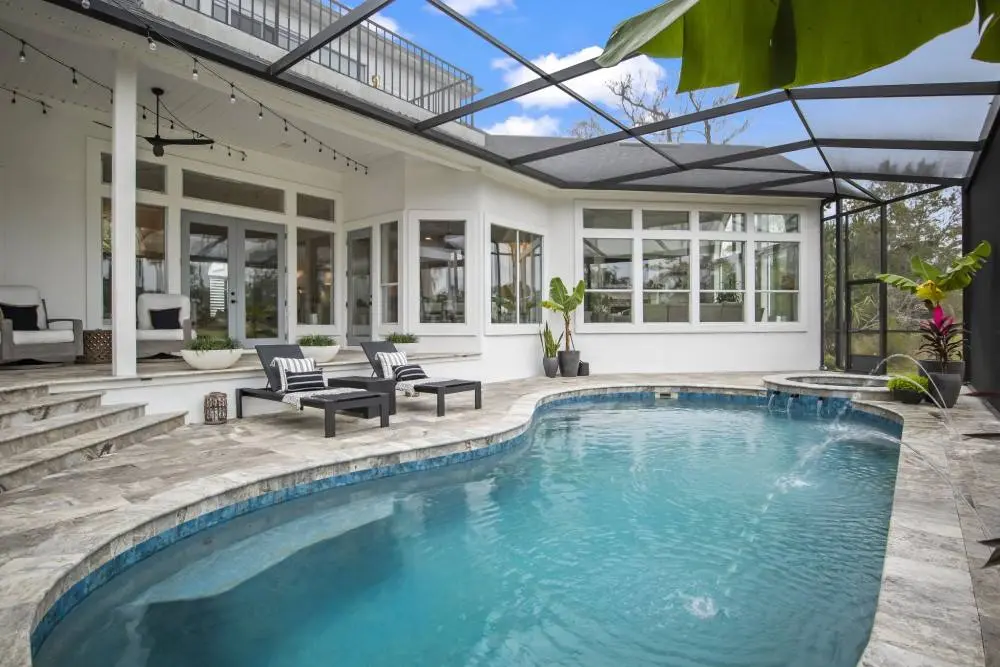Crystal-clear resort-style pool with blue tile accents, surrounded by travertine pavers, lounge chairs, and modern white poolhouse - Coastal Luxury Outdoors, Jacksonville, FL