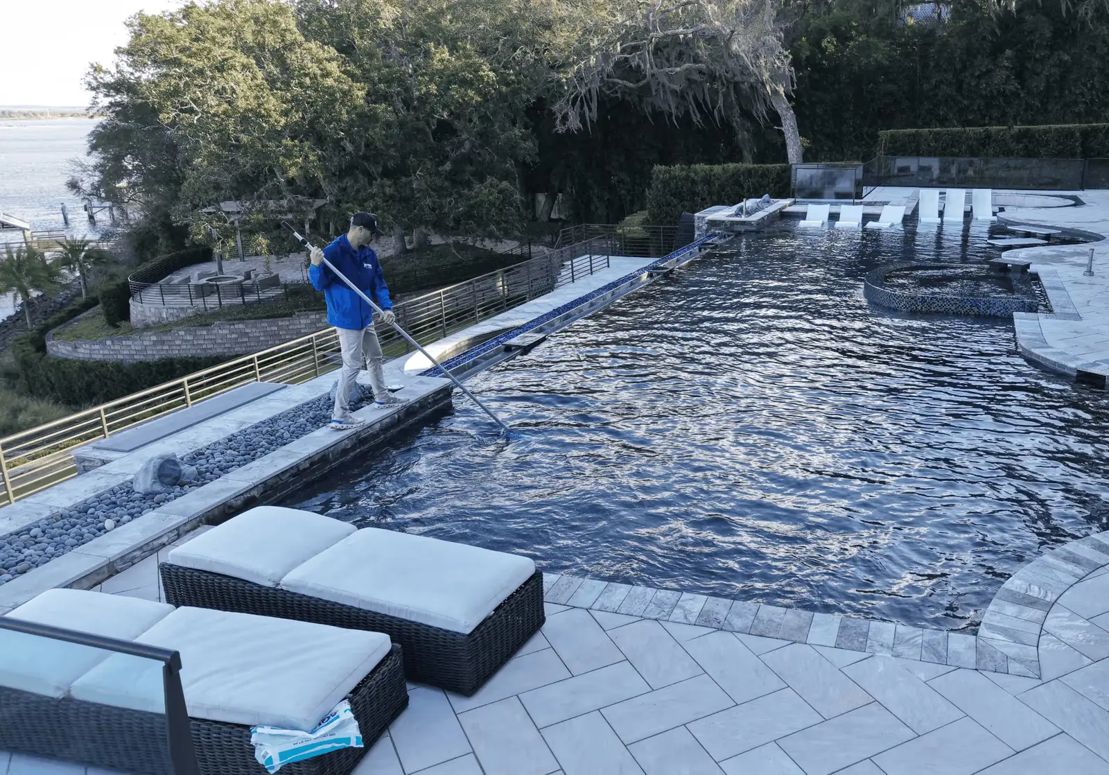 Technician in blue shirt using pool maintenance tool on clean blue pool with tile decking - Jomo Pool Service, Jacksonville, FL