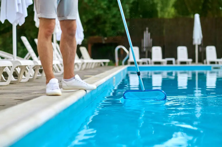 Technician using a blue pool skimmer on a bright, clean swimming pool with lounge chairs beside it - North East Florida Pool Service, Jacksonville, FL