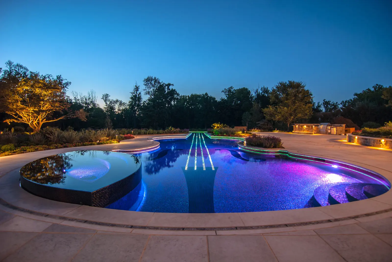 Luxurious swimming pool with vibrant blue and purple underwater lighting at dusk, surrounded by stone decking and illuminated landscaping - North East Florida Pool Service, Jacksonville, FL