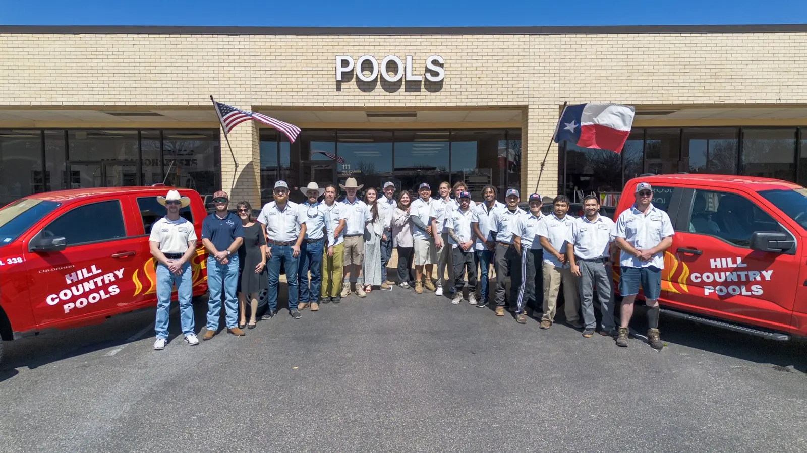 Hill Country Pools team in uniforms standing between branded red service trucks in front of their office - Hill Country Pools, Austin, TX
