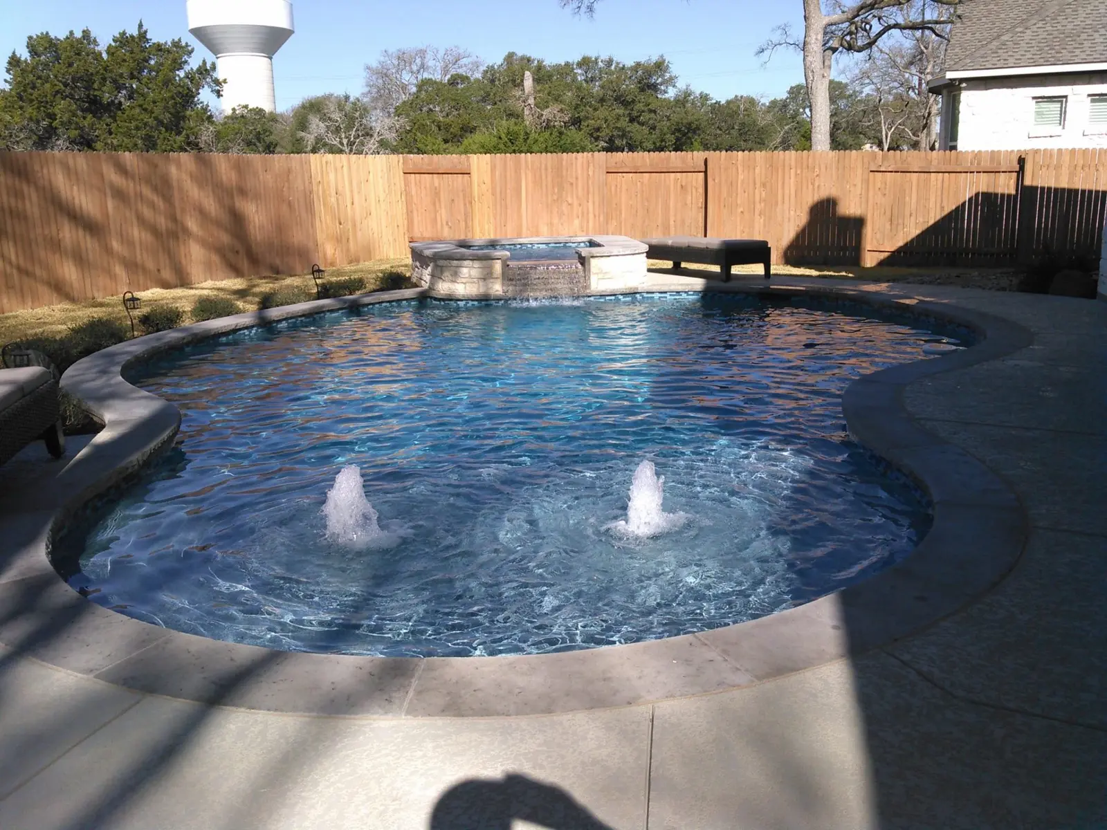 A sparkling clean backyard swimming pool with fountains and attached spa, surrounded by concrete deck and wooden fence - Prime Pool Service, Austin, TX