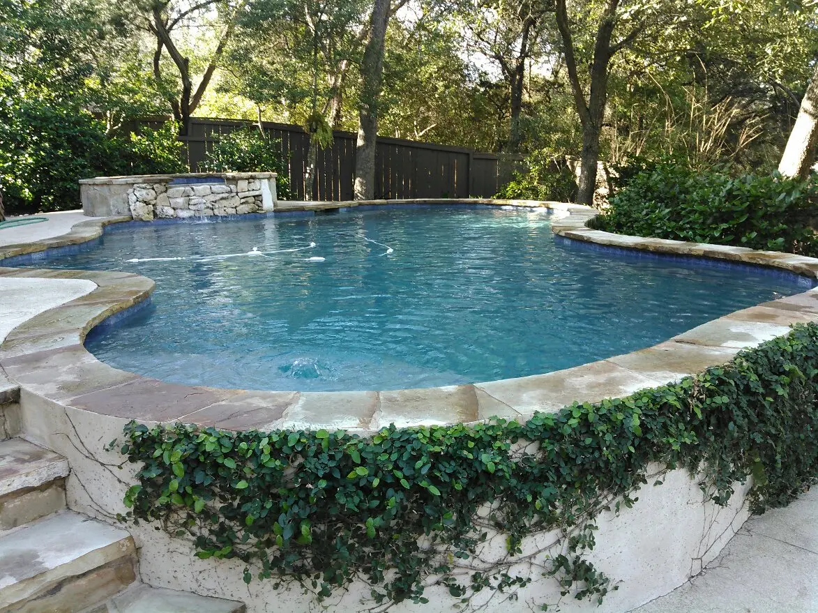 Sparkling blue swimming pool with stone deck and spa feature surrounded by green landscaping - Prime Pool Service, Austin, TX