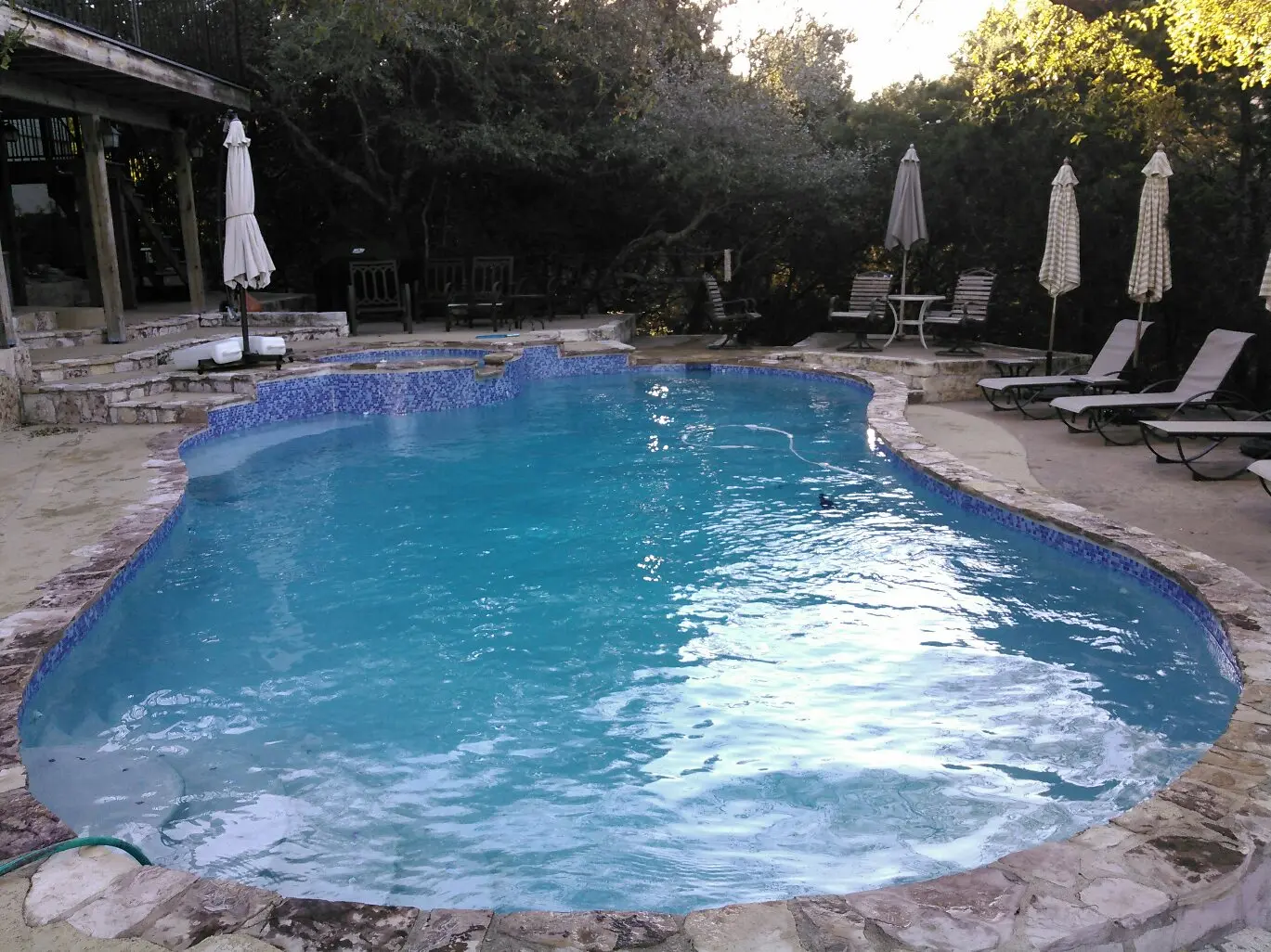 A sparkling blue resort-style pool surrounded by lounge chairs, umbrellas, and mature trees with stone patio - Prime Pool Service, Austin, TX