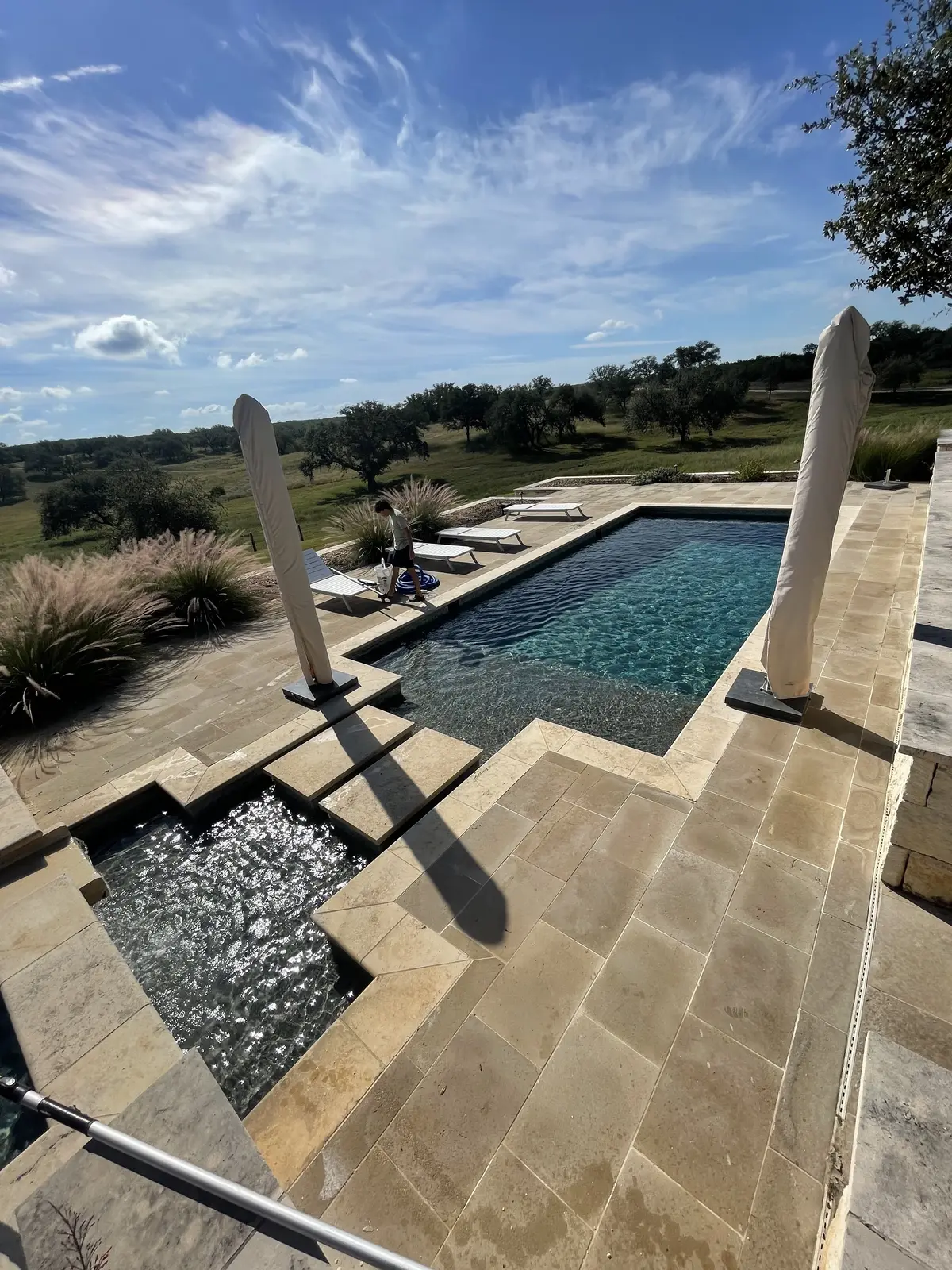 Sparkling blue pool with stone deck, lounge chairs, and umbrellas overlooking rolling landscape - Ranch Hand Pool Services, Corpus Christi, TX