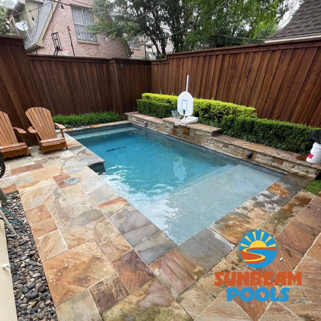 Sparkling clean backyard pool with stone patio, lounge chairs, and hedging beside wooden fence - Sunbeam Pools, Dallas, TX