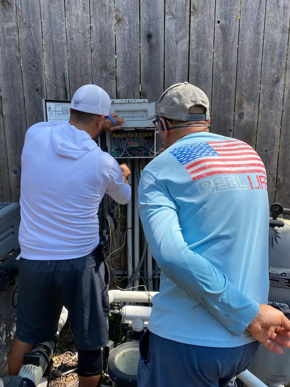 Two pool technicians in branded shirts servicing pool equipment control system against wooden fence - Elkin's Pool Service, Fort Worth, TX