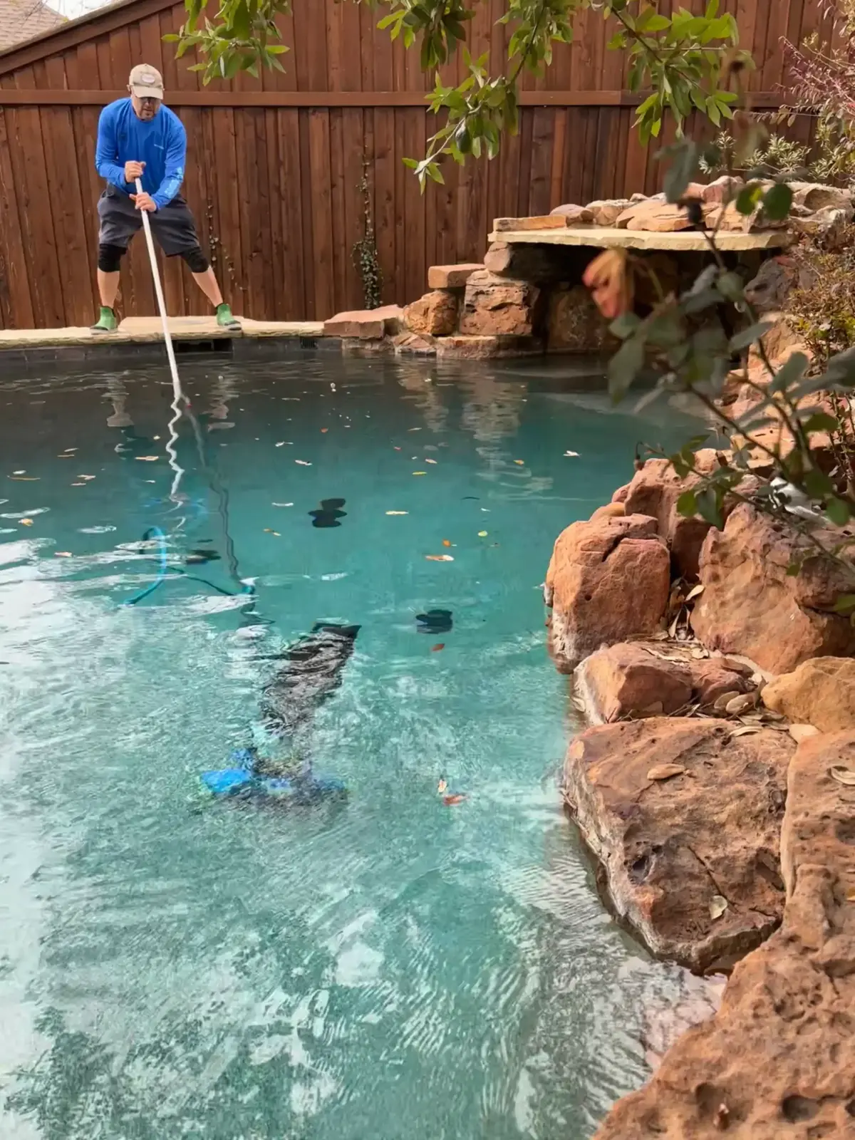 Technician using pool skimmer net to clean sparkling turquoise pool with stone waterfall feature - Elkin's Pool Service, Fort Worth, TX