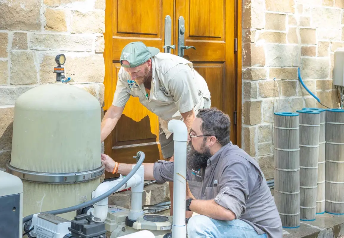 Two technicians service pool equipment including filters and a large tank at a residential home - Manning Pool Service, Houston, TX