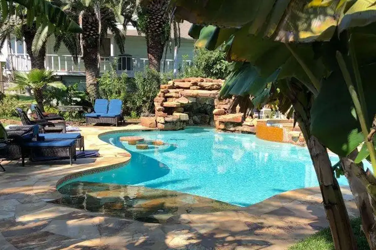 Crystal-clear blue swimming pool with rock waterfall feature, lounge chairs, and lush tropical landscaping - Manning Pool Service, Houston, TX